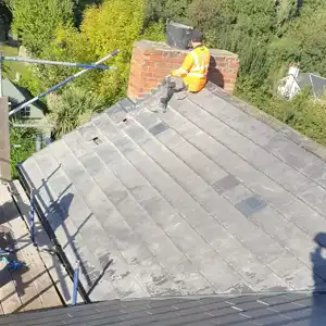 A worker in a bright orange vest is sitting on a sloped roof, repairing chimney bricks, with greenery and buildings visible in the background.